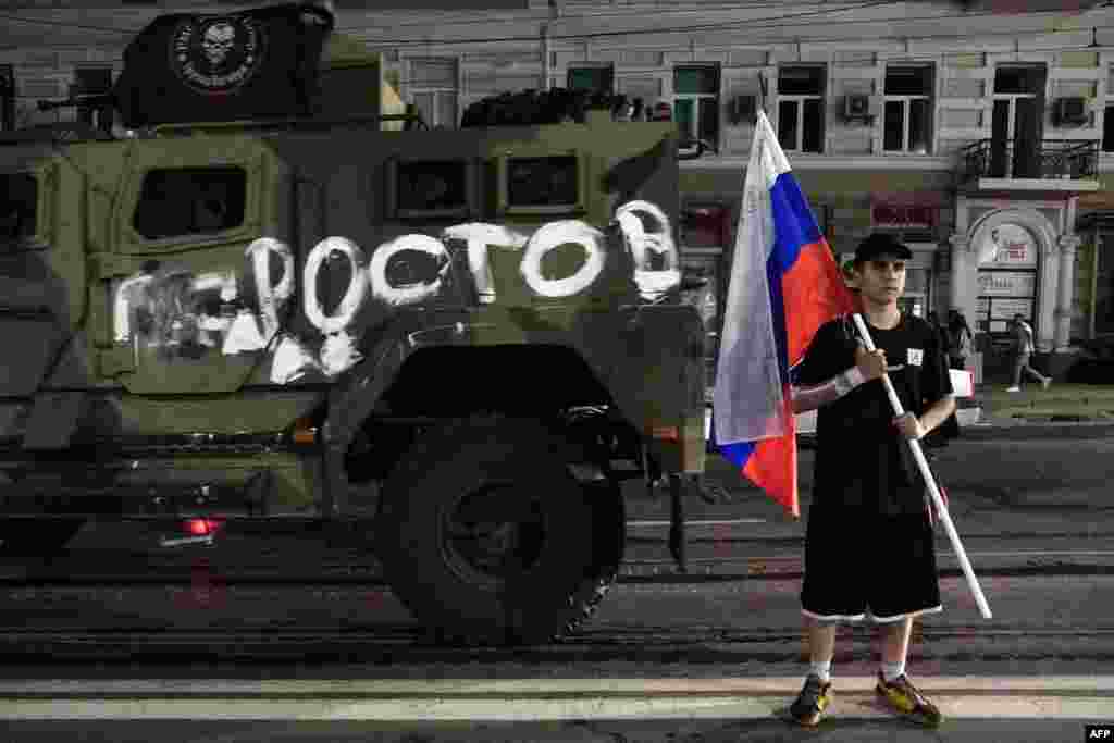 A man holds the Russian national flag in front of a Wagner vehicle with a painting that reads "Rostov." 