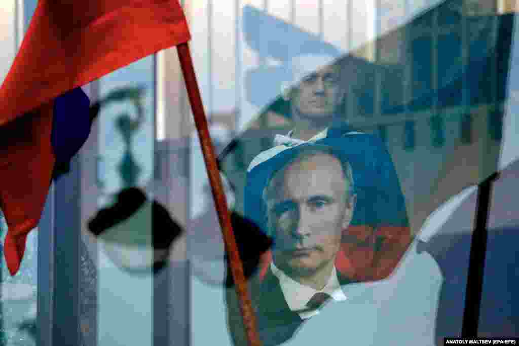 Мужчина в одежде с изображением президента России Владимира Путина смотрит в окно, когда полиция охраняет территорию вокруг концертного зала «Октябрьский» в Санкт-Петербурге 