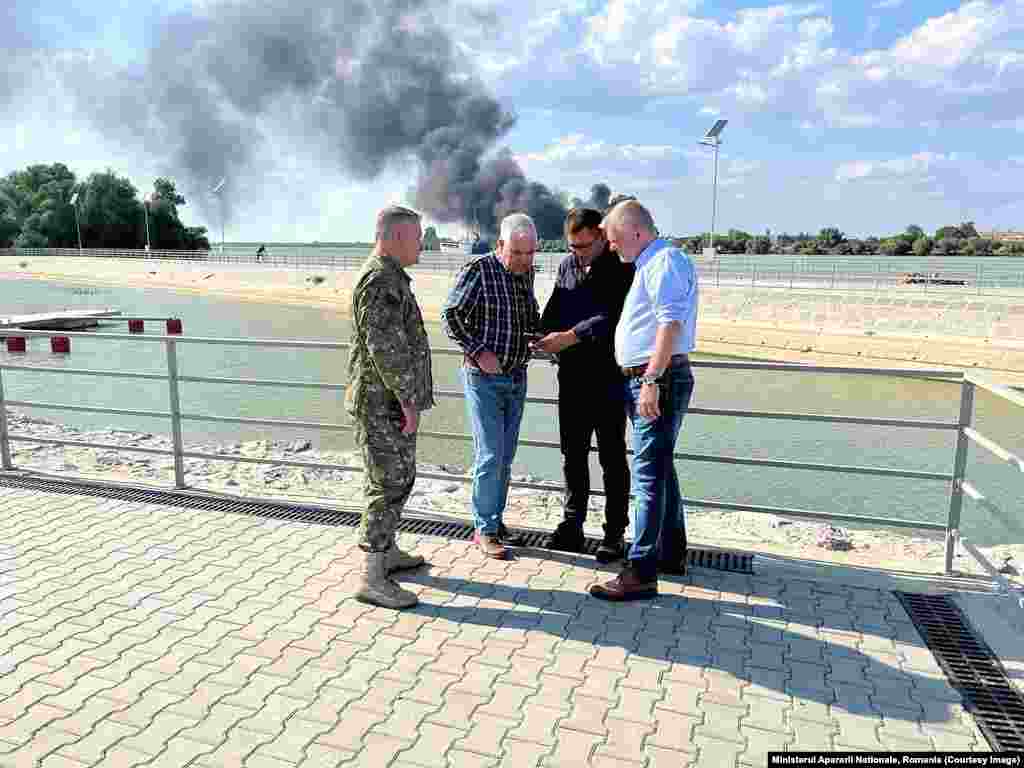 Smoke rises from Ukrainian territory as Romania’s Defense Minister Angel Tilvar (in plaid shirt) consults with other men in an image released by the ministry on September 6. In a September 6 Facebook post, Romania’s Defense Ministry appeared to contradict the assertions of the president, announcing it had discovered “elements that resemble drone remains” on Romanian territory just across the border from Ukraine's Izmayil, which it vowed to investigate.