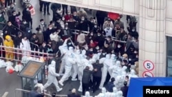 Şanhaýdaky öýleriň biriniň ýaşaýjylary daşaryk çykmaga rugsat bermeýän gorag eşiklerini geýen polisiýa işgärleri bilen garpyşýarlar. 2022-nji ýylyň 30-njy noýabry