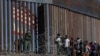 Migranți din țări din America Centrală se uită prin gardul de la frontiera cu Mexic, lângă punctul de trecere El Chaparral, Baja, California